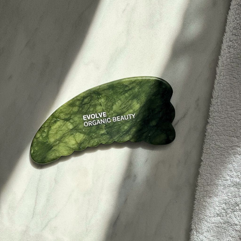 green jade body gua sha on marble table with sun shining on it and white towel