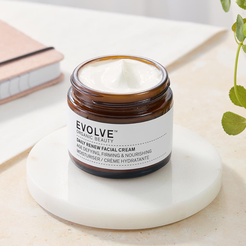 Open amber glass jar of Daily Renew Facial Cream on a marble coaster. Surrounded by folliage and a notebook in a lifestyle setting.