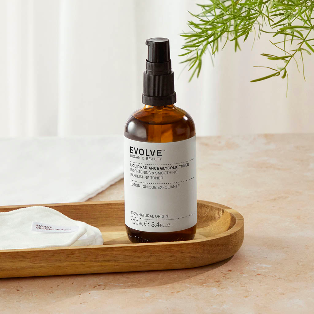 Liquid Radiance Glycolic Toner bottle on top of a wooden trey with some Reusable Bamboo Makeup pads next to it all on top of beige counter with green leaves in the corner