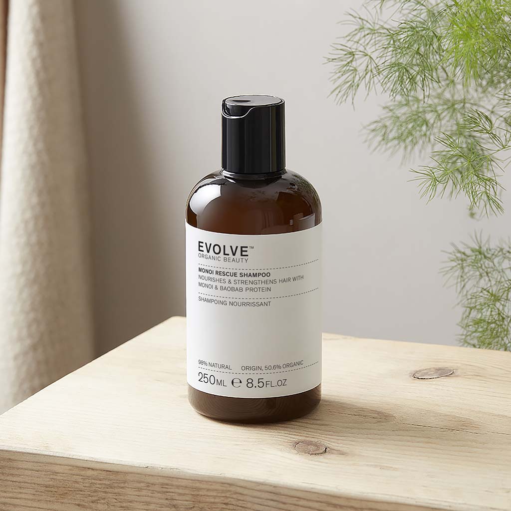 Bottle of Monoi Rescue Shampoo on a natural pale wood shelf with a plain neutral wall behind, hanging fern plant and natural coloured curtain
