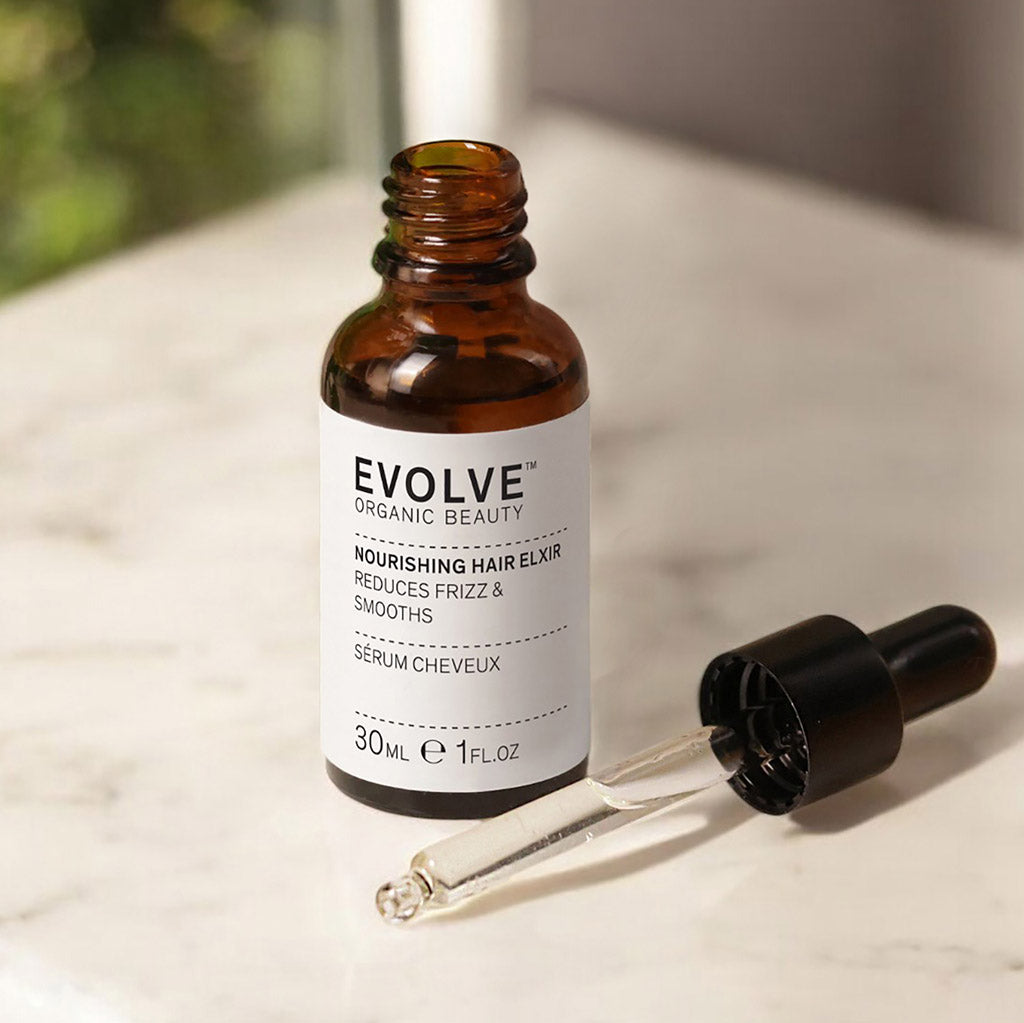 An open bottle of Nourishing Hair Elixir with the glass dropper lying down in front of the bottle on a white marble surface with a window in the background showing blurred greenery outside.