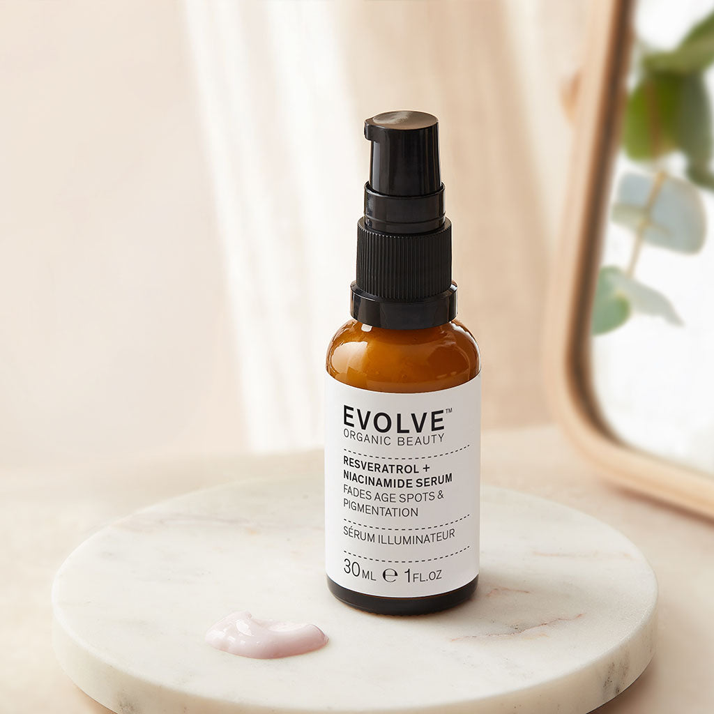 bottle of resveratrol + niacinamide serum on top of a marble coaster with a light pink swatch of the product next to it with mirror in soft focus on the side