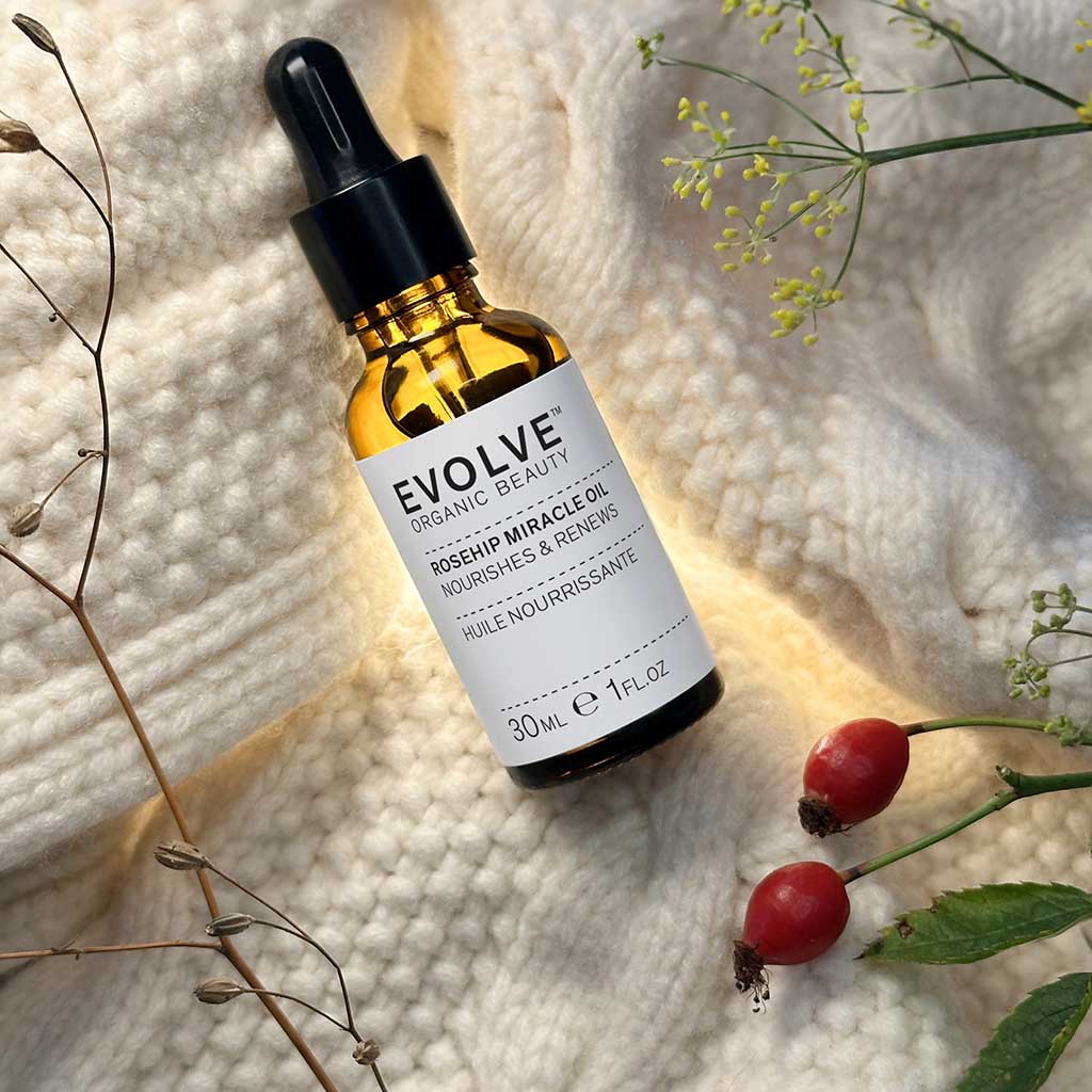 a bottle of rosehip miracle oil on top of white soft knitted blanket with rosehips and other stems on the side