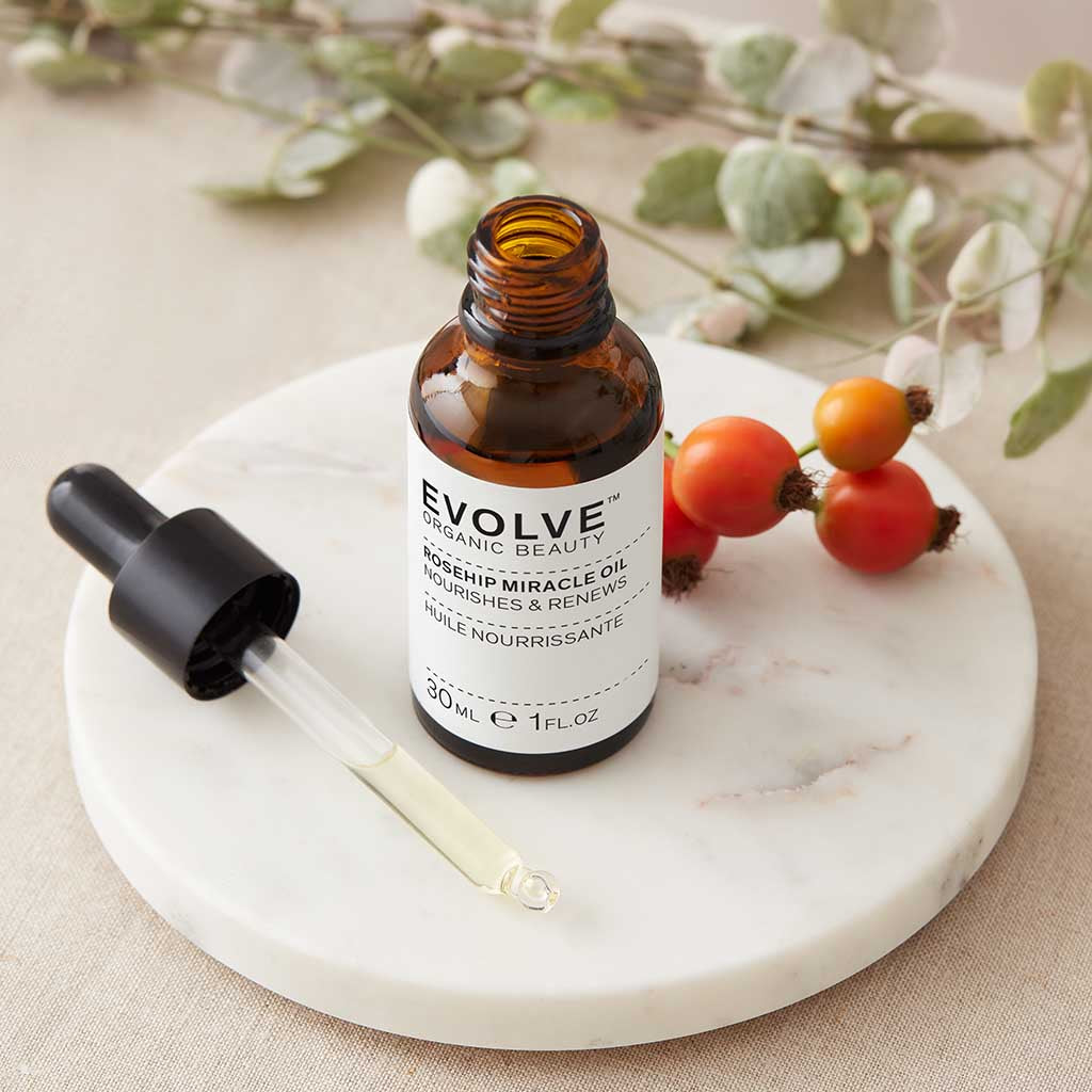 an open bottle of rosehip miracle oil on top of marble coaster with rosehips and pipette filled with the oil next to it and some foliage on the side