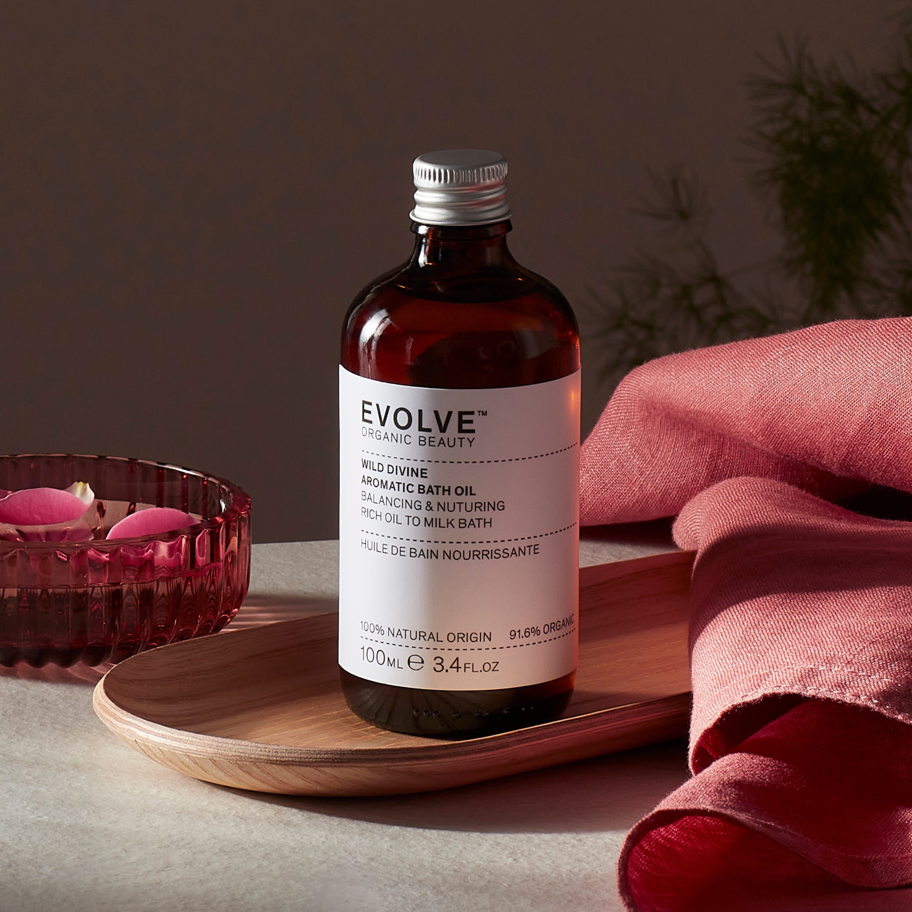 Evolve organic beauty Wild Divine Aromatic Bath Oil in Brown bottle with cap on a wooden tray with dark pink towel and rose petals in bowl on a dark beige background