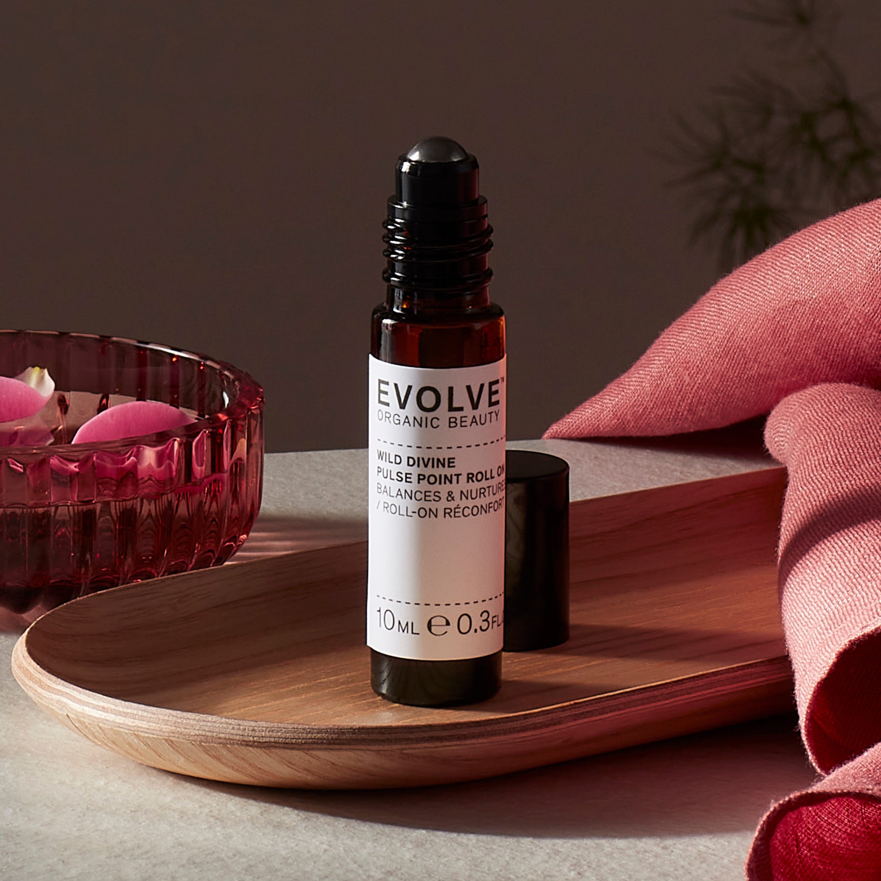 Evolve organic beauty Wild Divine Pulse Point Roll On in Brown bottle with cap on a wooden tray with dark pink towel and rose petals in bowl on a dark beige background