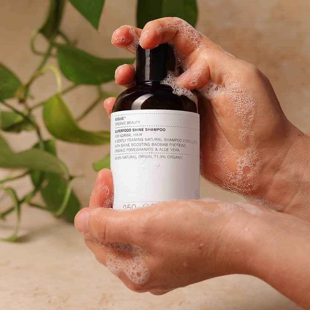 Hands holding a bottle of Superfood Shine Shampoo with the soft white foam of the shampoo around the hands and bottle. In the background is a green plan in soft focus with neutral coloured walls