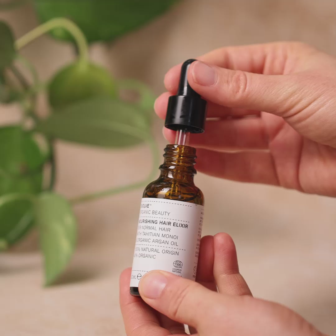 Video of hands holding a bottle of Nourishing Hair Elixir, opening it up and showing the Elixir texture of oil dropping out of the bottle. A green plant is in soft focus in the background.