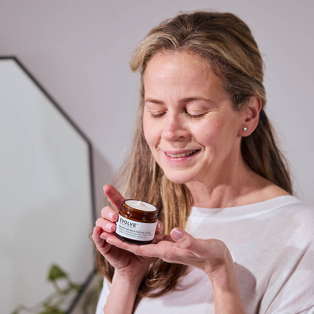 model holding Age Defying Multi Peptide Cream jar in hands and smiling while looking at it, in a bathroom setting with mirror and foliage