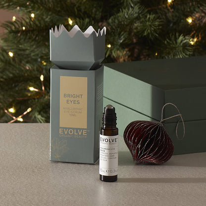 Evolve skincare hyaluronic eye serum with christmas packaging and decorative ornament on a surface with a Christmas tree in the background