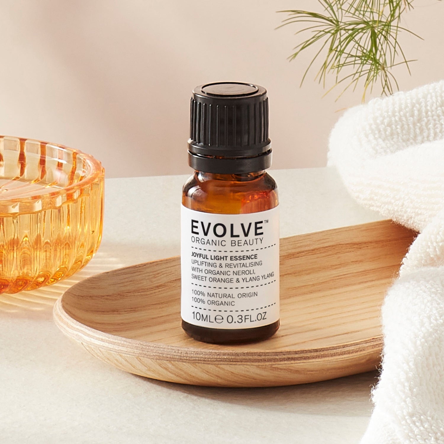 Evolve organic beauty Joyful Light Essence in Brown bottle with cap on a wooden tray with towel on a beige background