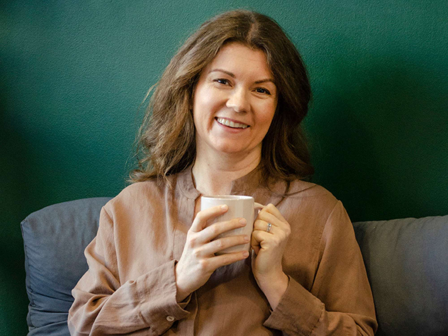 Laura Rudoe founder of Evolve Organic Beauty sitting in front of dark green wall holding a mug and smiling warmly at camera