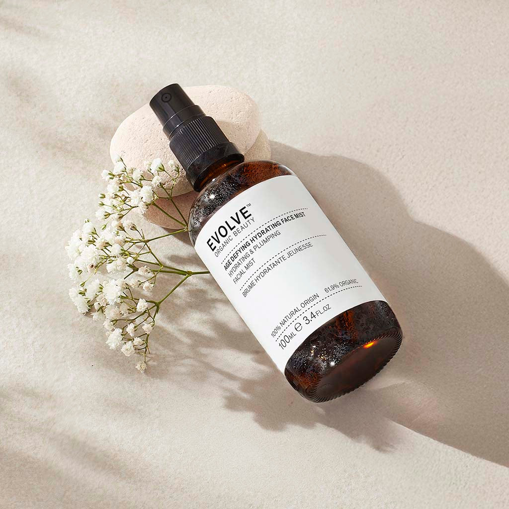 misted bottle of age defying hydrating face mist on top of two pebbles with white bud flower stem under it