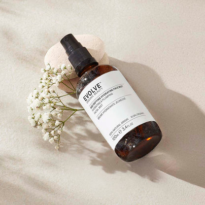 misted bottle of age defying hydrating face mist on top of two pebbles with white bud flower stem under it