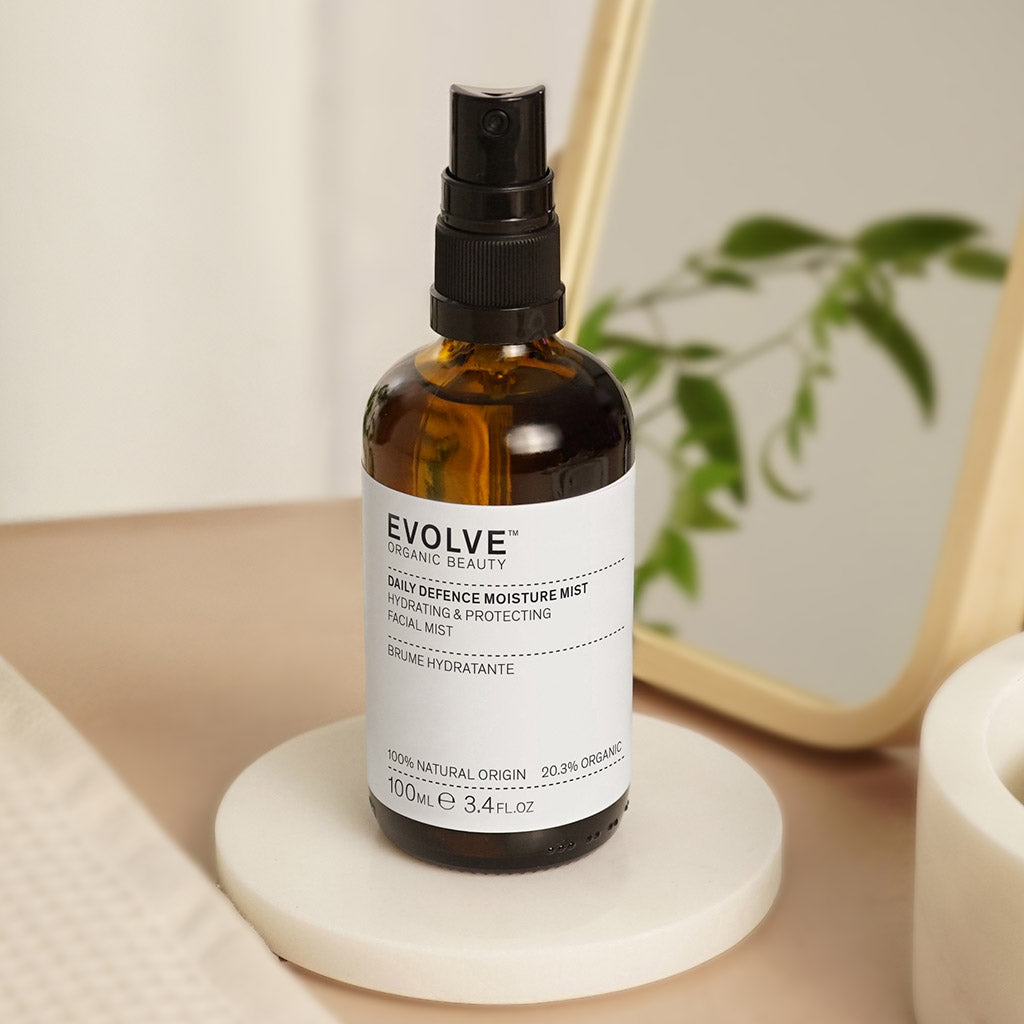 bottle of daily defence moisture mist on top of white coaster with a towel on the side and wooden framed mirror with foliage in the background