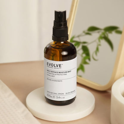 bottle of daily defence moisture mist on top of white coaster with a towel on the side and wooden framed mirror with foliage in the background