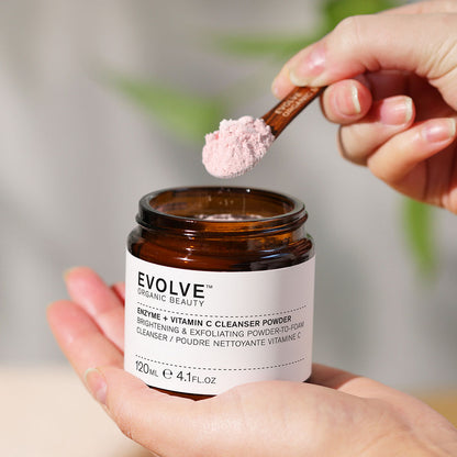 a jar of enzyme + vitamin C powder cleanser held in one hand with other hand picking the powder cleanser up with a small wooden spoon
