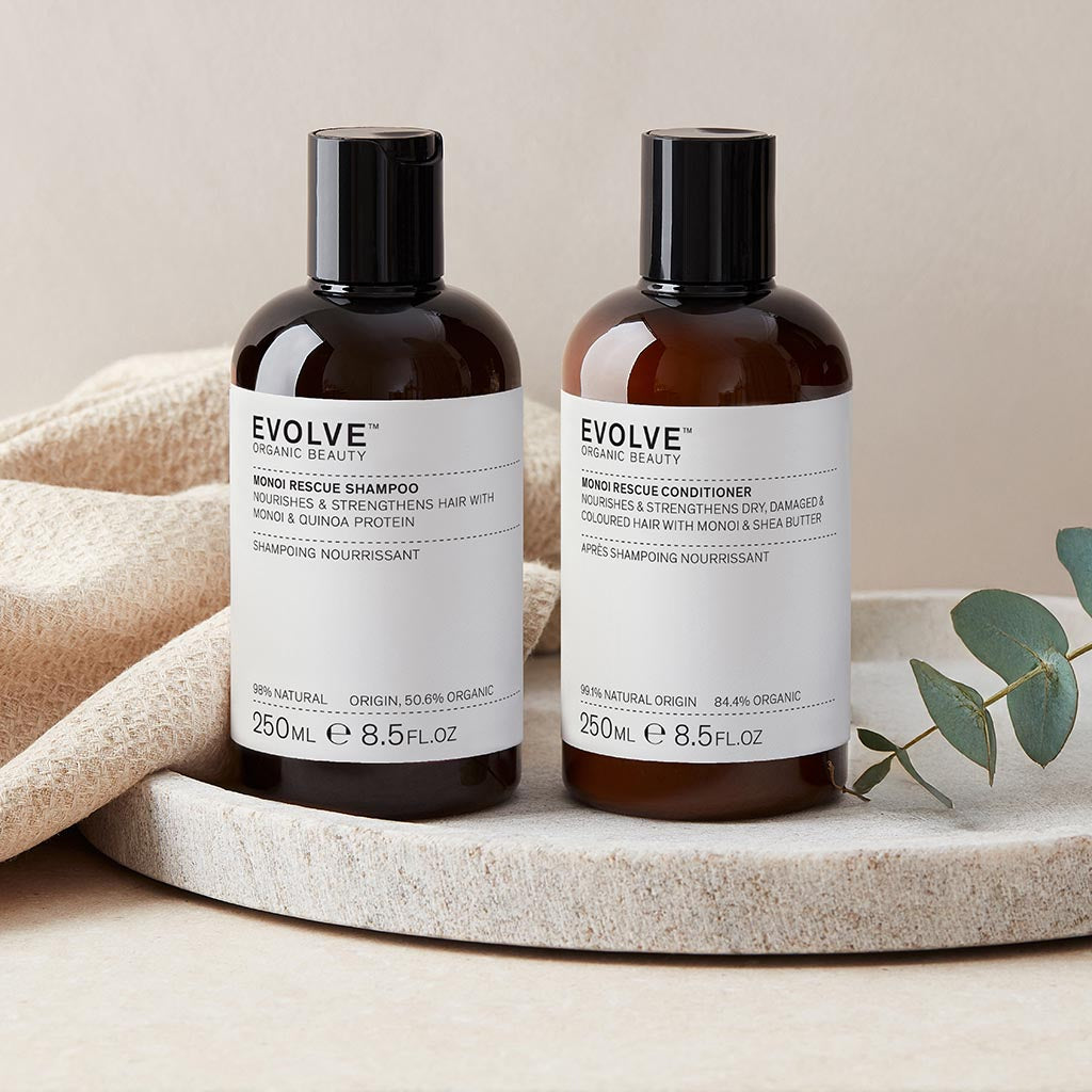 Bottle of Monoi Rescue Shampoo and Conditioner on a natural stone tray with a plain neutral wall behind, eucalyptus branch and neutral towel