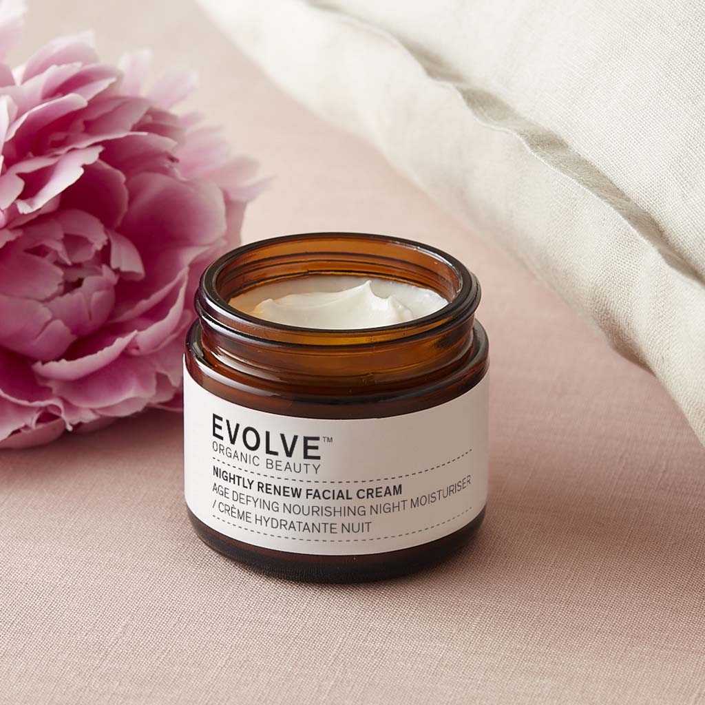 Open amber glass jar of Nightly Renew Facial Cream on a pale pink bed with a pink peony and pillow behind it