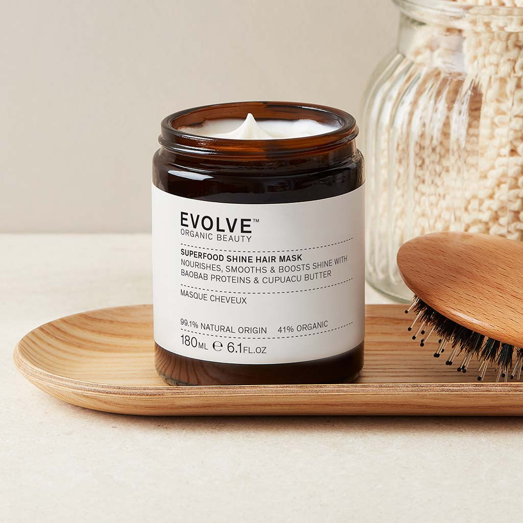 Open jar of Superfood Shine Hair Mask on a small bamboo tray with a natural wooden hair brush, on a natural stone counter and wall behind. In the background there is a clear ribbed glass jar filled with a natural washcloth