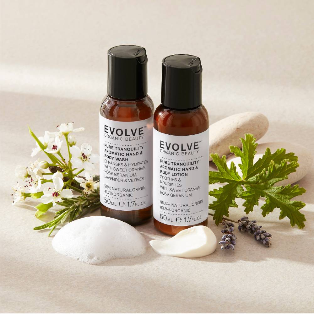 bottle of pure tranquility aromatic wash and  lotion travel sizes on beige background  with rosemary and geranium leaves, lavander and stones with swatches showing each product&