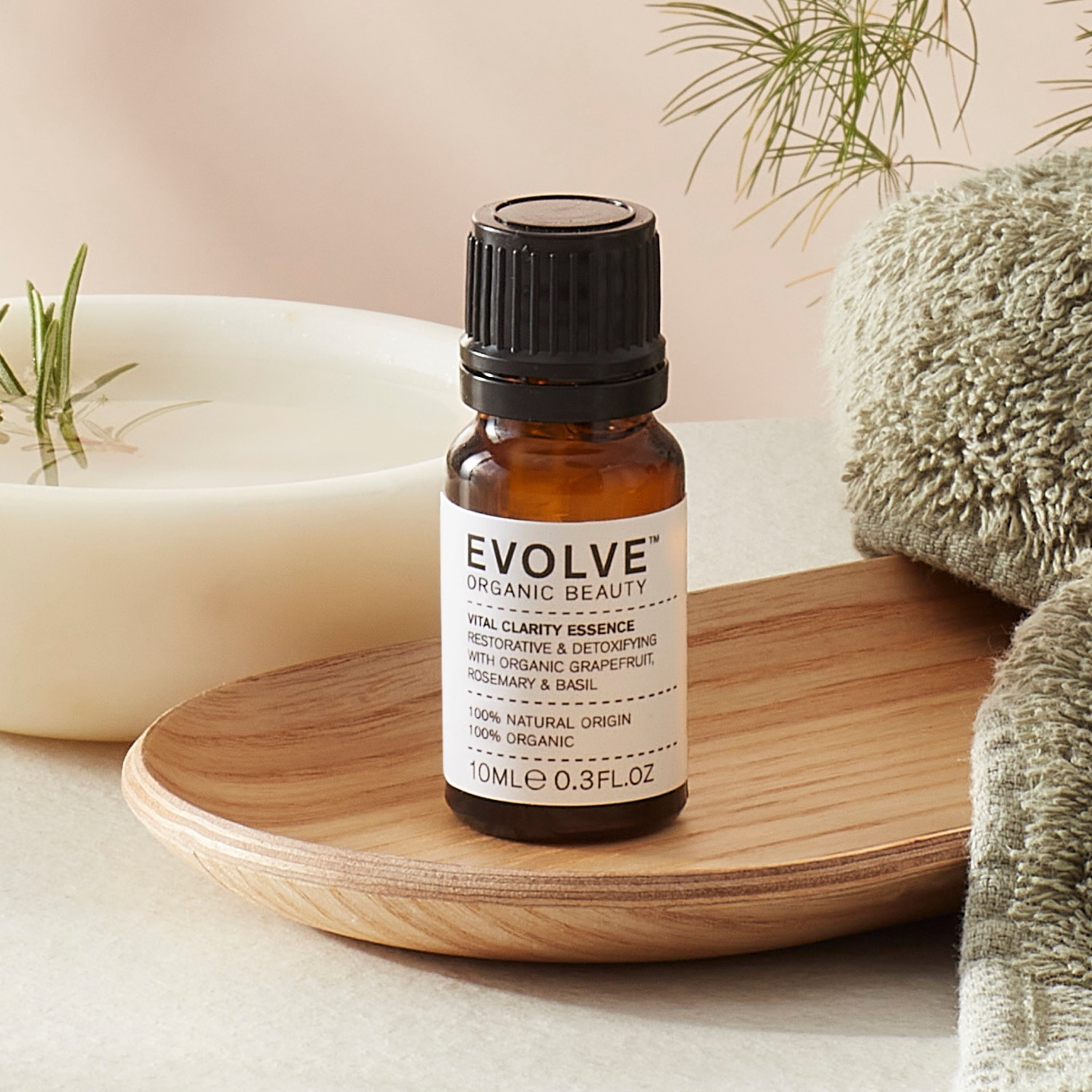 Evolve organic beauty Vital Clarity Essence in Brown bottle with cap on a wooden tray with green towel on a beige background