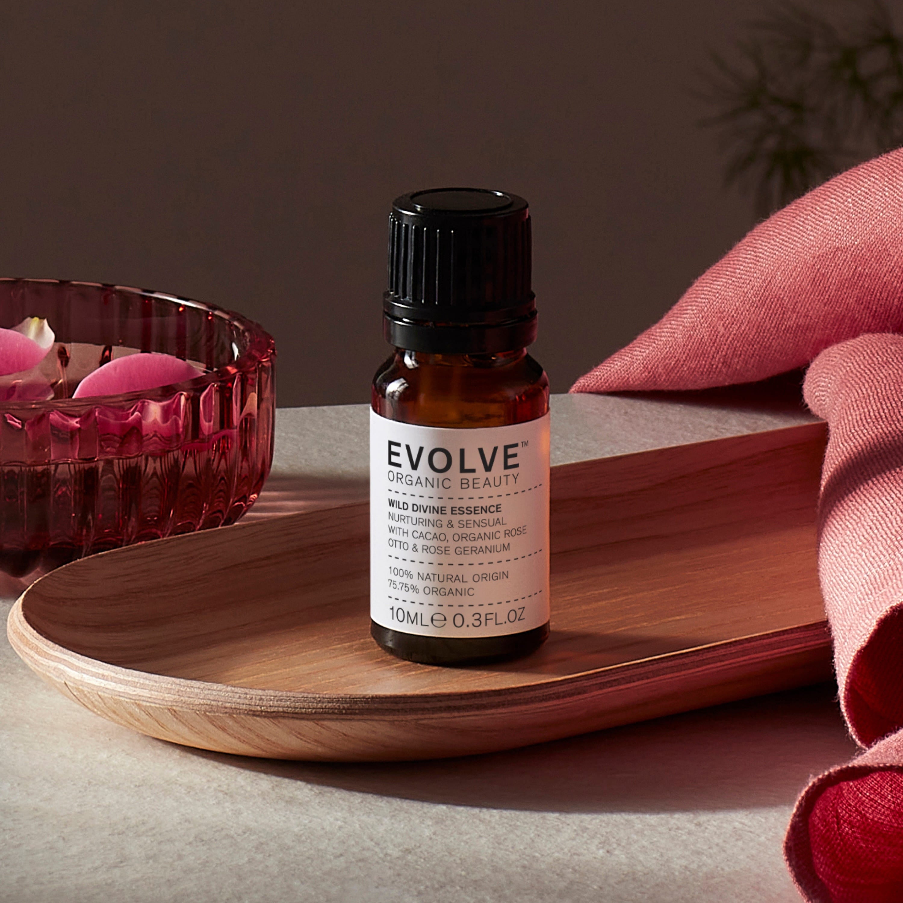Evolve organic beauty Wild Divine Essence in Brown bottle with cap on a wooden tray with dark pink towel and rose petals in bowl on a dark beige background
