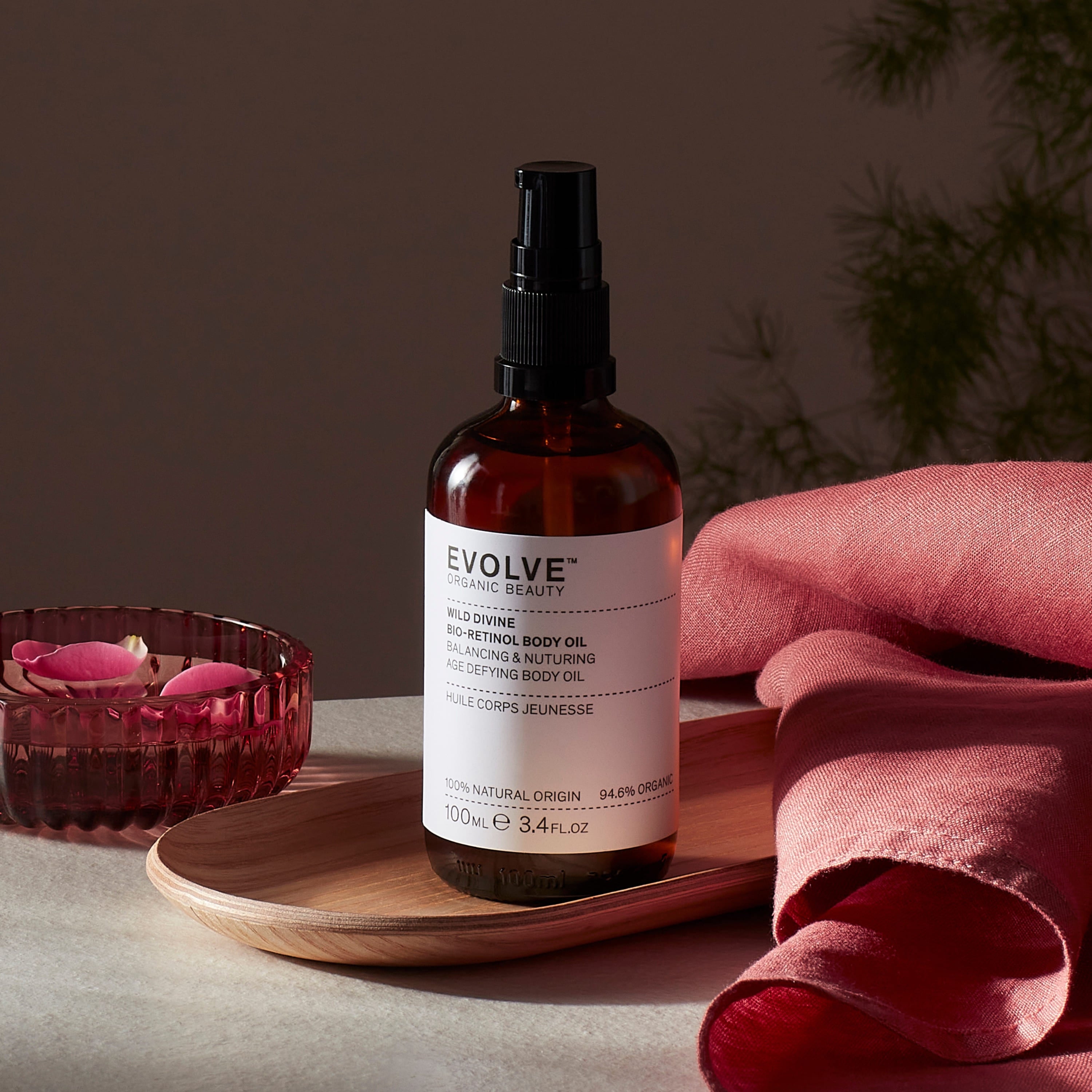Evolve organic beauty Wild Divine Aromatic Body Oil  in Brown bottle with pump on a wooden tray with dark pink towel and rose petals in bowl on a dark beige background