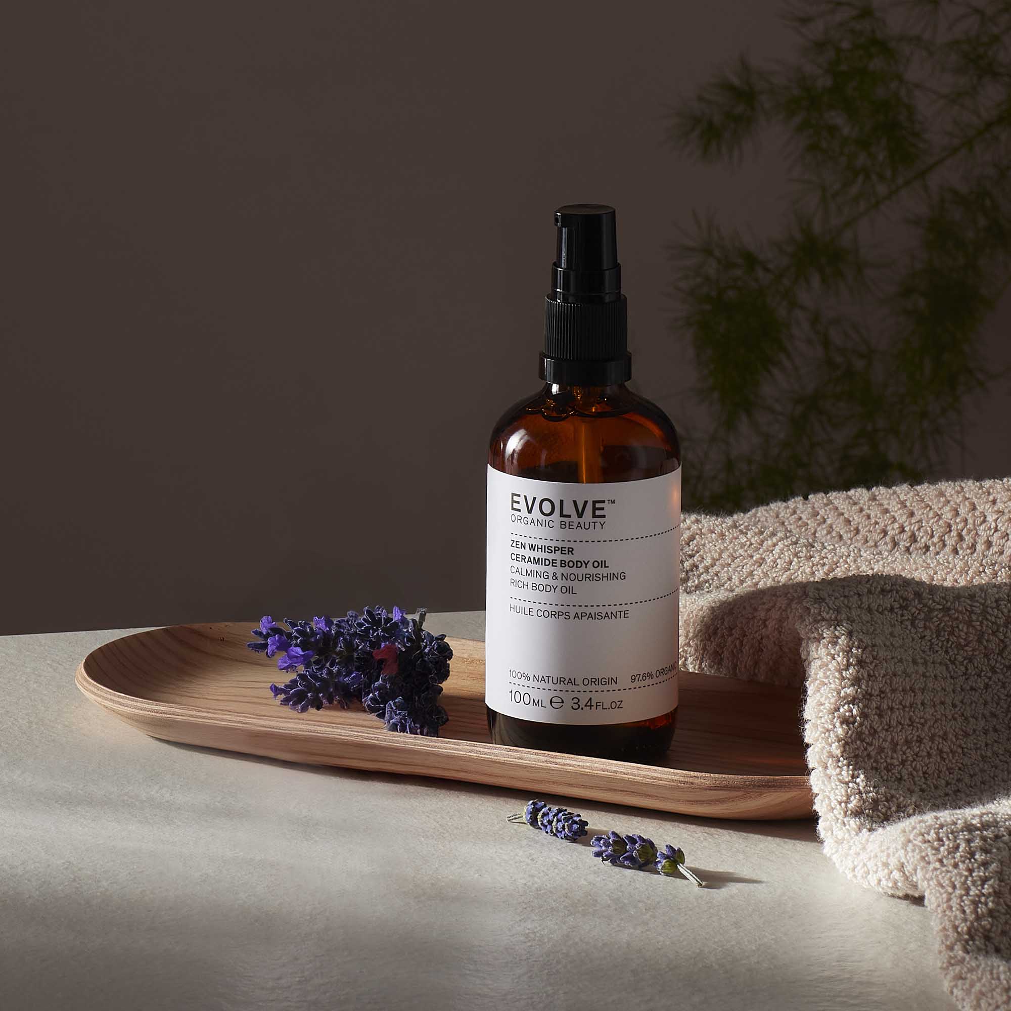 Evolve organic beauty Zen Whisper Ceramide Body Oil in Brown bottle with cap on a wooden tray with beige towel  on a dark beige background with lavender