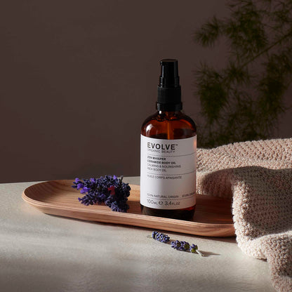 Evolve organic beauty Zen Whisper Ceramide Body Oil in Brown bottle with cap on a wooden tray with beige towel  on a dark beige background with lavender