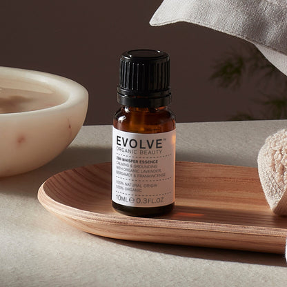 Evolve organic beauty Zen Whisper Essence  in Brown bottle with cap on a wooden tray with beige towel  on a dark beige background