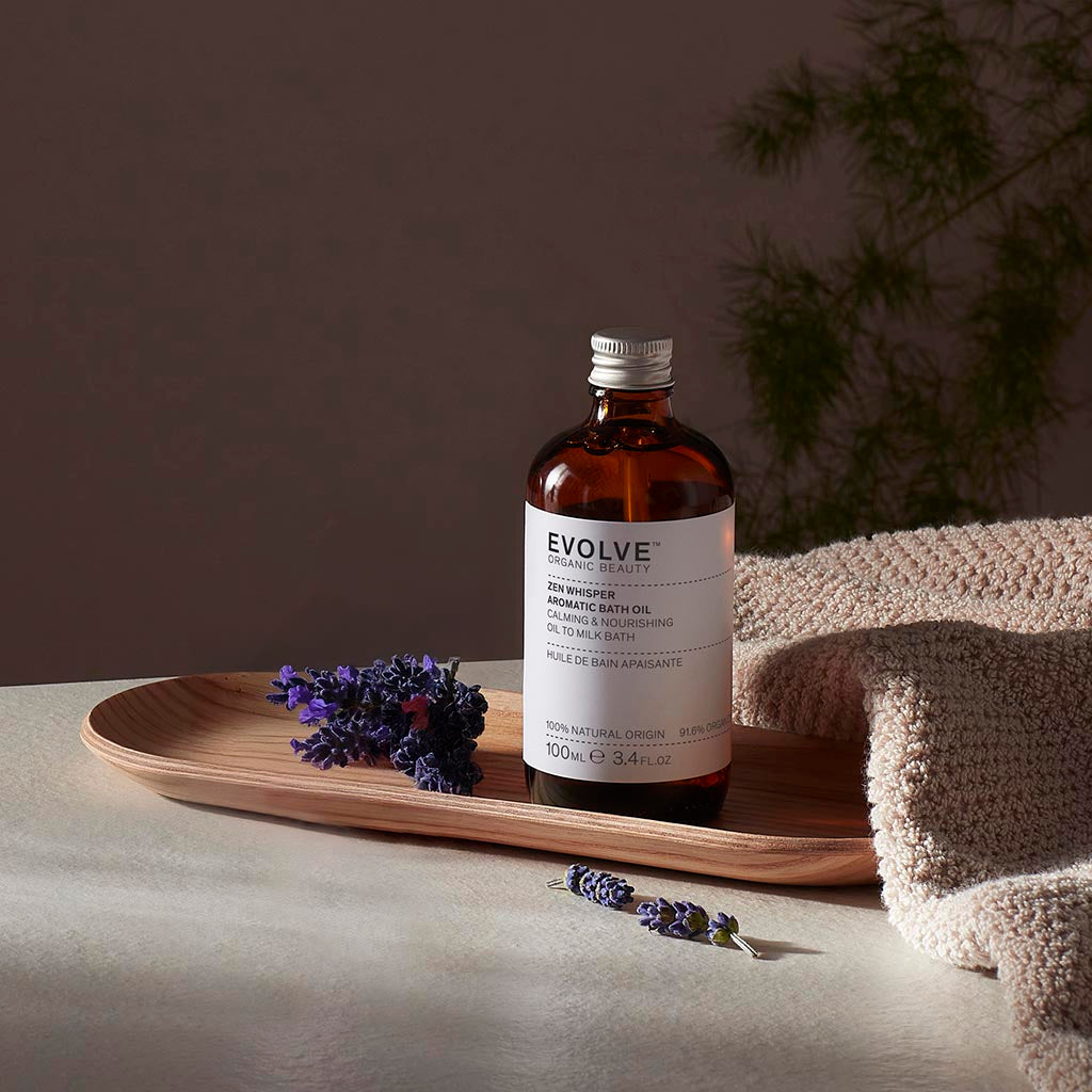 Evolve organic beauty Zen Whisper Ceramide Bath Oil in Brown bottle with cap on a wooden tray with beige towel  on a dark beige background with lavender