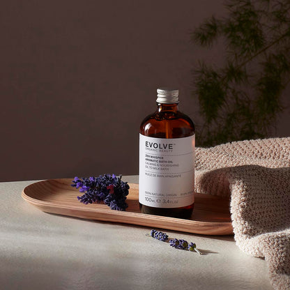 Evolve organic beauty Zen Whisper Ceramide Bath Oil in Brown bottle with cap on a wooden tray with beige towel  on a dark beige background with lavender