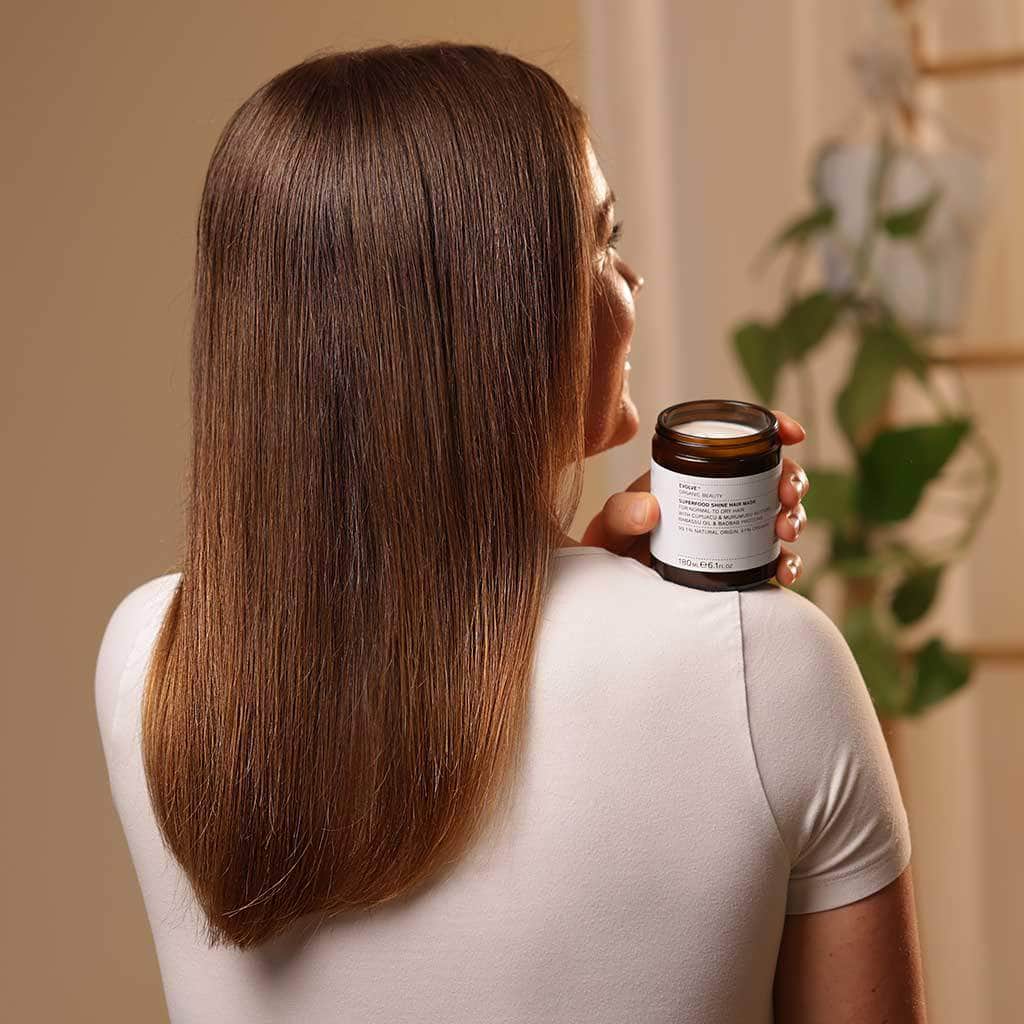 Back view of a woman with long silky shiny straight hair holding an open jar of Superfood Shine Hair Mask on her shoulder, wearing a white T-shirt. Background is warm beige with a soft-focus hanging plant in the distance.